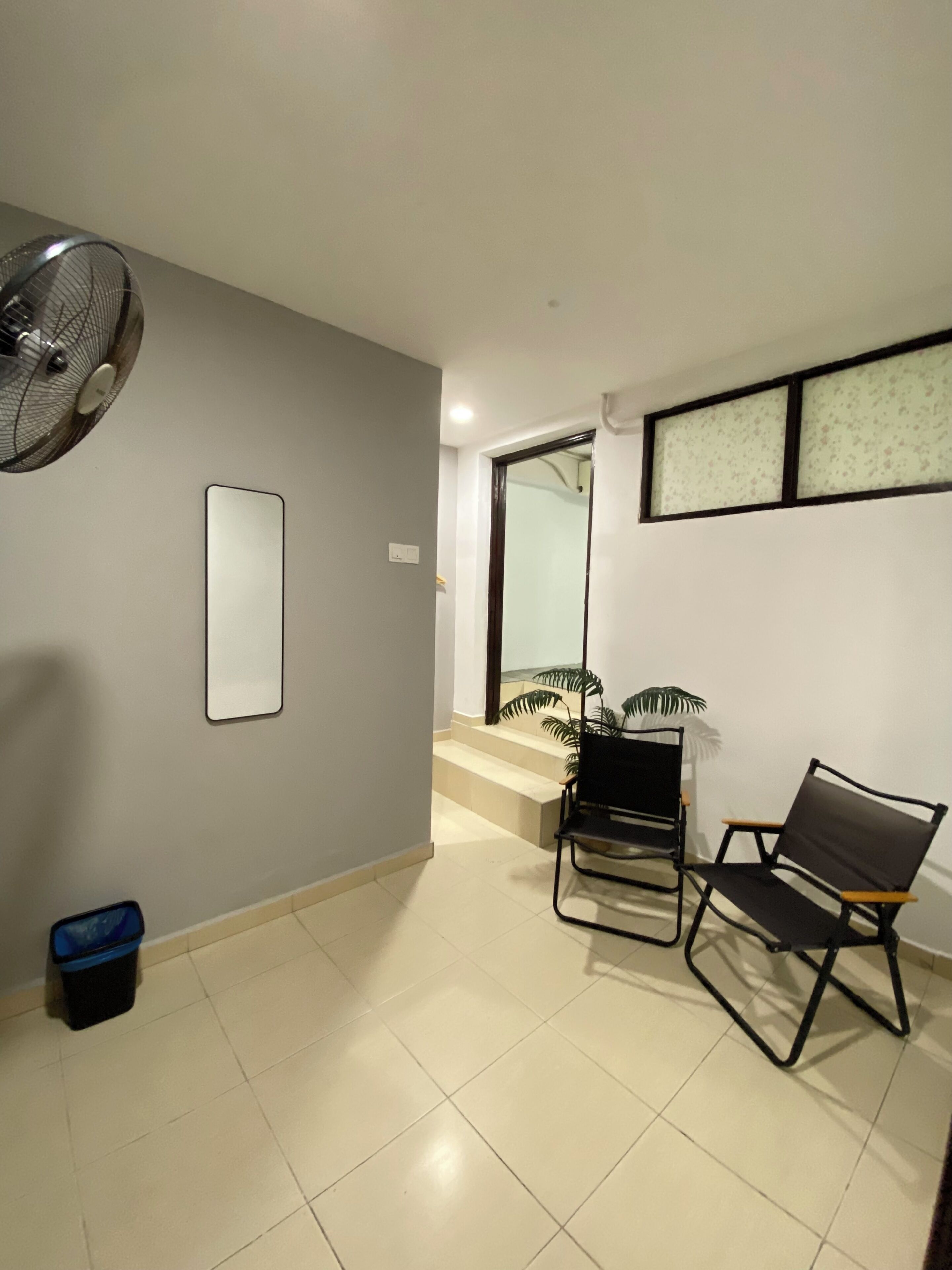 Una habitación con ventilador, una silla, un pequeño bote de basura y una escalera que lleva a otra zona.