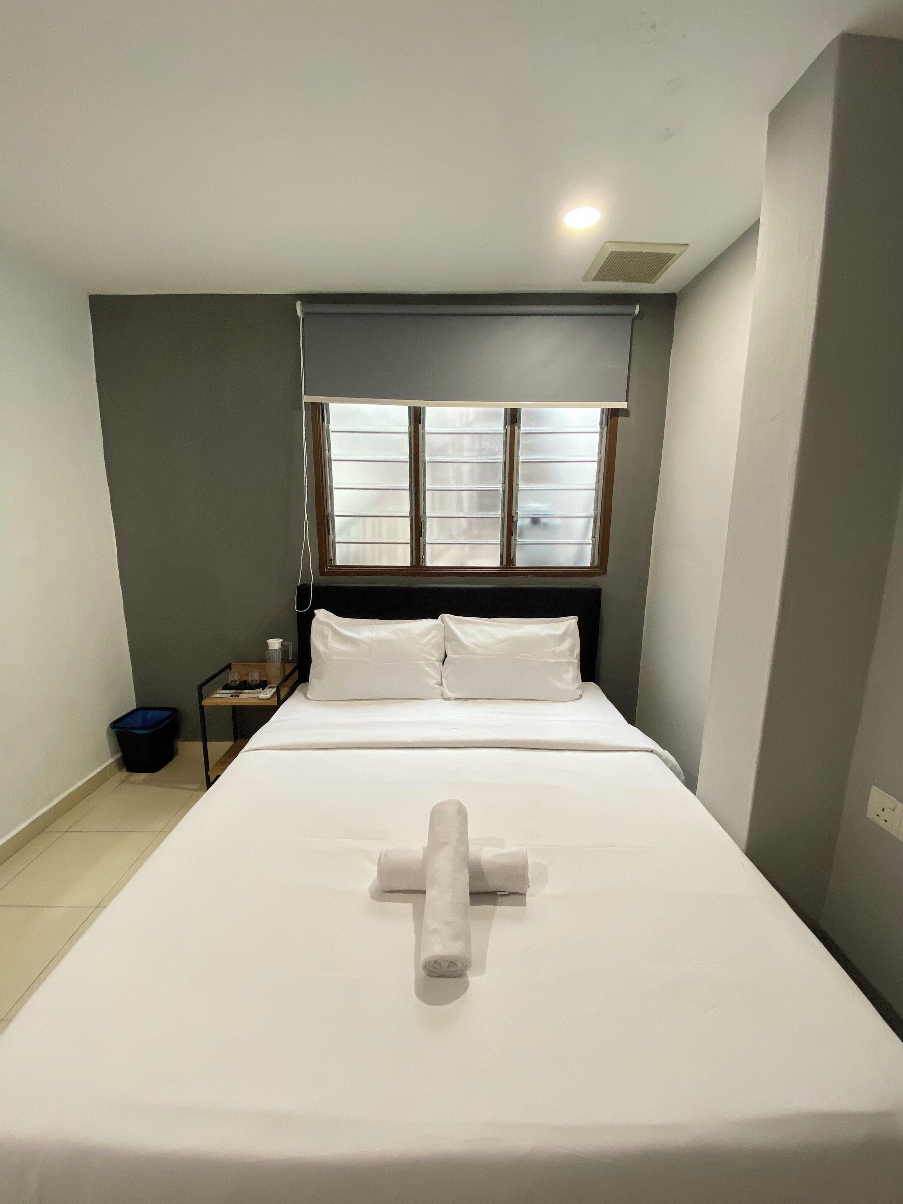 A neatly made bed with a rolled towel in the center, a bedside table with a lamp, and a window with blinds.