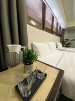 Habitación de hotel con cama, mesita de noche con jarrón, vasos y bandeja, y una planta en maceta.