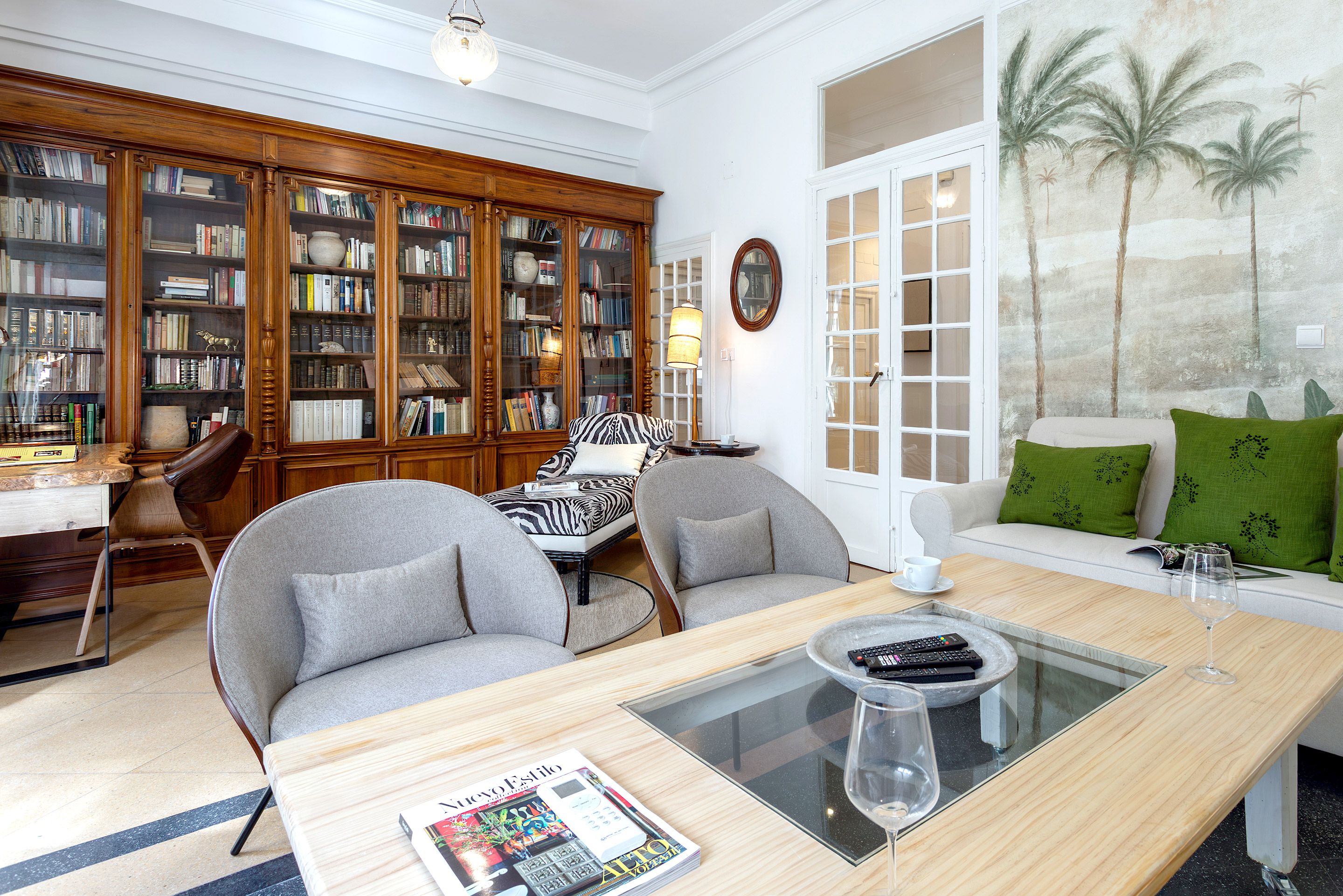 A living room with a sofa, armchairs, a coffee table, a bookshelf, and a wall with a palm tree mural.