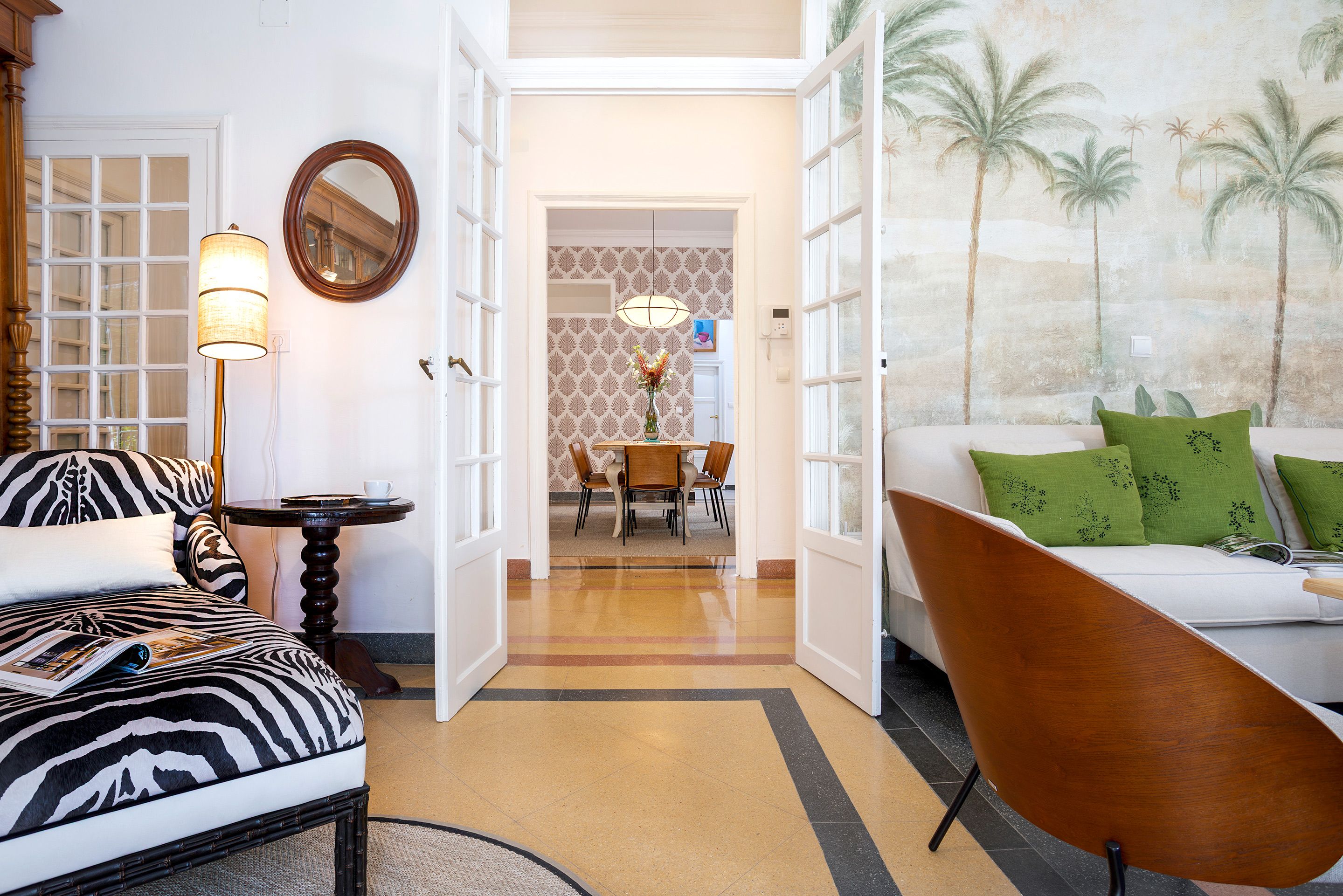 A living room with a zebra-patterned sofa, a wooden chair, a table with a lamp, and a painting of palm trees.