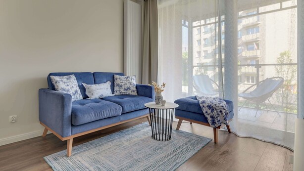 A modern living room with a blue sofa, a small round table, and a view of a building through large windows. - Scala 16 | Parking | City Center (Gdańsk)