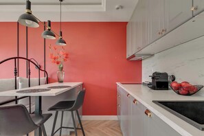 A compact kitchen with a red accent wall, a dining area with a white table and black chairs, and a coffee machine on the counter. - Rezydencja Wintera 1A | River View (Gdańsk)