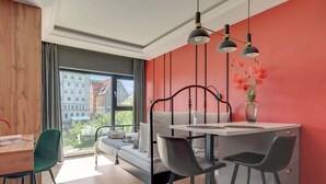 A modern bedroom with a bed, a dining table, two chairs, a window with a view of buildings, and a red wall. - Rezydencja Wintera 1A | River View (Gdańsk)