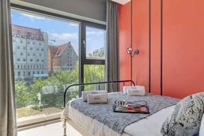 A bedroom with a large window, a bed with a grey blanket, and a red wall. - Rezydencja Wintera 1A | River View (Gdańsk)
