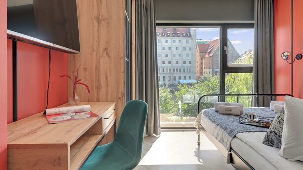 A modern hotel room with a bed, a desk, a chair, a TV, and a balcony with a city view. - Rezydencja Wintera 1A | River View (Gdańsk)