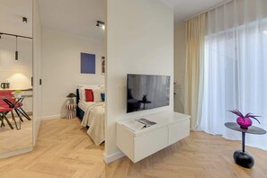 Ein modernes Wohnzimmer mit Flachbildfernseher, einer weißen Unterhaltungseinheit, einem Essbereich mit Tisch und Stühlen sowie einem großen Fenster mit durchsichtigen Vorhängen.