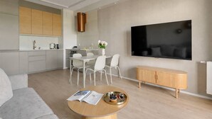 Ein modernes Wohnzimmer mit einem grauen Sofa, einem Couchtisch mit Zeitschriften, einem Essbereich mit weißem Tisch und Stühlen, einer Küche mit Holzschränken und einem an der Wand befestigten Fernseher.