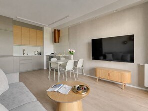 Ein modernes Wohnzimmer mit einem grauen Sofa, einem Couchtisch mit Zeitschriften, einem Essbereich mit weißem Tisch und Stühlen, einer Küche mit Holzschränken und einem an der Wand befestigten Fernseher.
