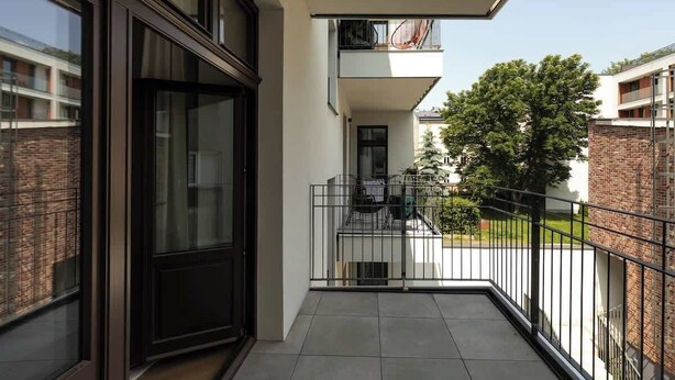 Un balcón con barandilla de metal, puerta de vidrio y vista a un árbol y edificios vecinos.