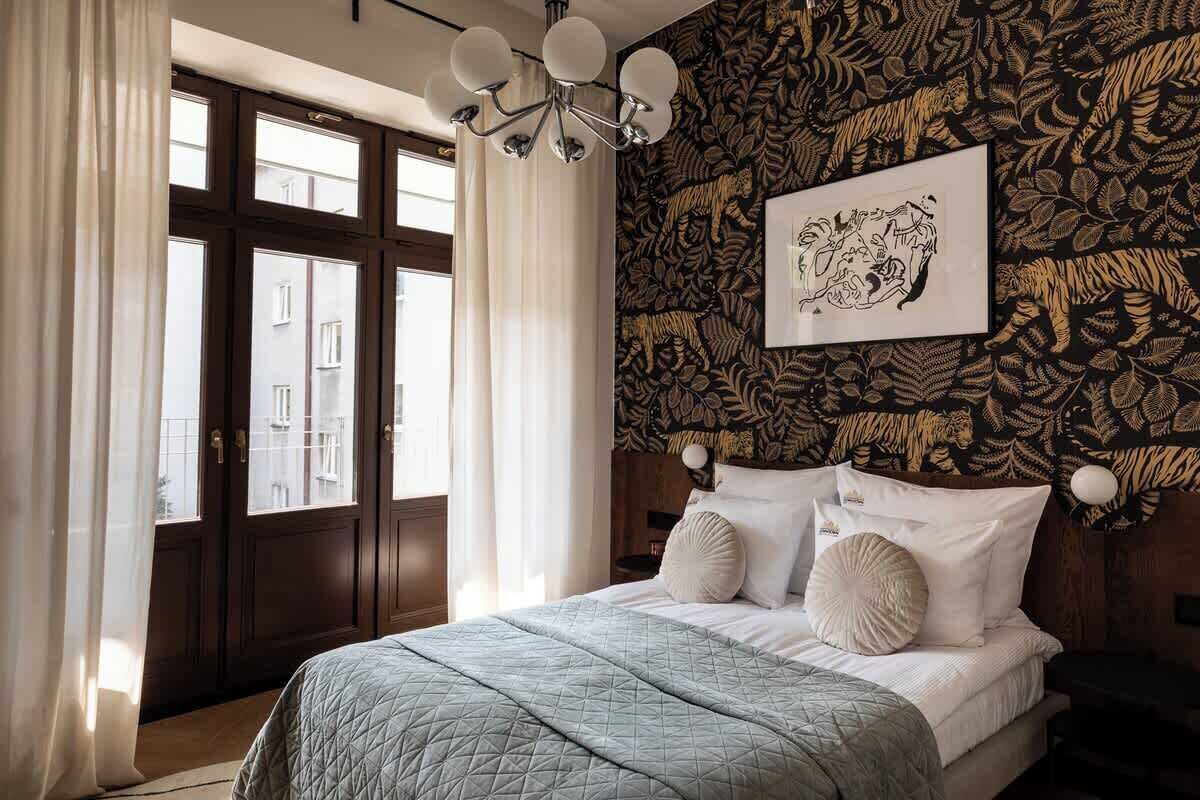 A bedroom with a bed, curtains, a window, a wall with a leaf pattern, and a chandelier.