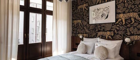 A bedroom with a bed, curtains, a window, a wall with a leaf pattern, and a chandelier.