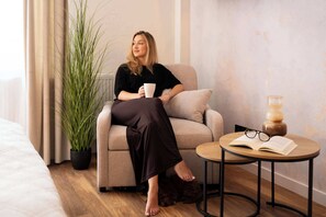 Eine Person sitzt auf einem Sofa in einem Zimmer mit einem Blumentopf, einem runden Tisch mit einem Buch und Brillen, und einem Bett im Hintergrund.