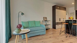 Una sala de estar moderna con un sofá, una mesita redonda con un florero, una lámpara de pie y una pequeña zona de kitchenette con un frutero.