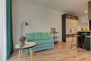 A modern living room with a sofa, a small round table with a vase, a floor lamp, and a kitchenette area with a fruit bowl. - Dwie Motławy 40 | Riverside | Balcony (Gdańsk)
