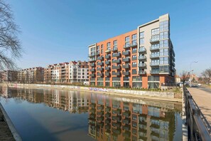 A modern apartment building with multiple balconies, a reflection in the water, and a clear sky. - Dwie Motławy 40 | Riverside | Balcony (Gdańsk)