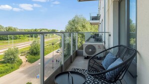 A balcony with a glass railing, a black wicker chair, a small round table, and a view of a railway track and greenery. - Letnicka 31 with Balcony | Perfect Stay (Gdańsk)
