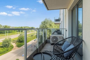 Ein Balkon mit Glasgeländer, einem schwarzen Korbstuhl, einem kleinen runden Tisch und Blick auf Gleise und Grünflächen.