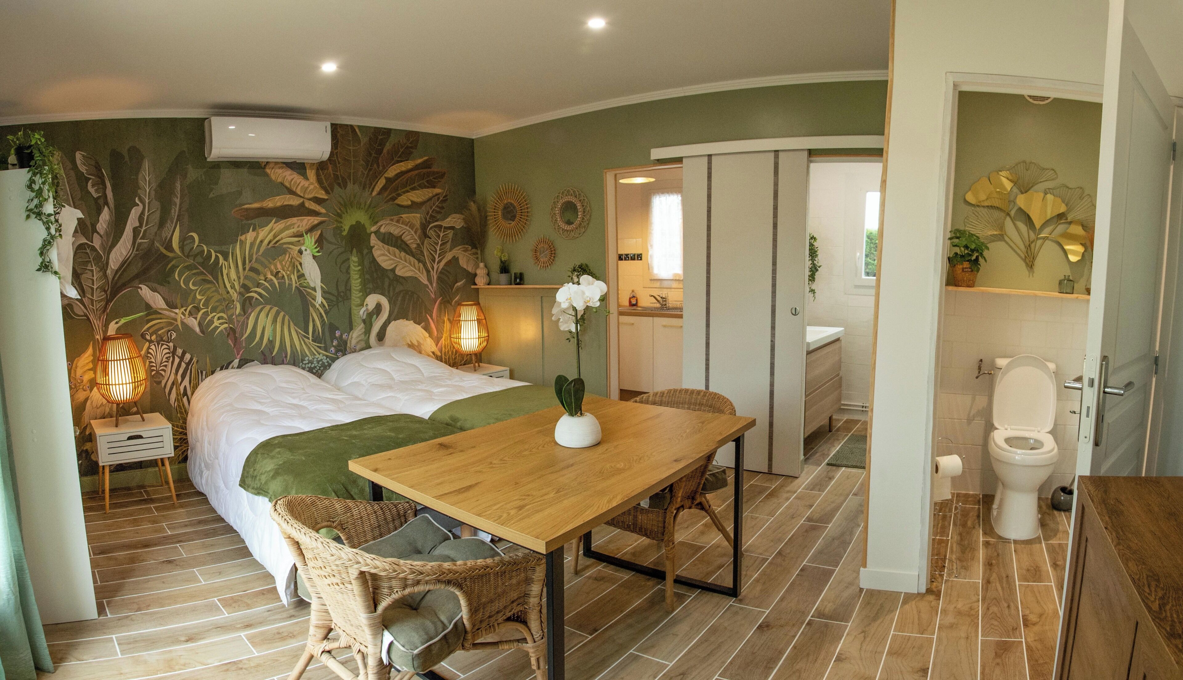 Quarto com uma cama, uma mesa com um vaso de flores, uma cadeira de vime e vista para o banheiro.