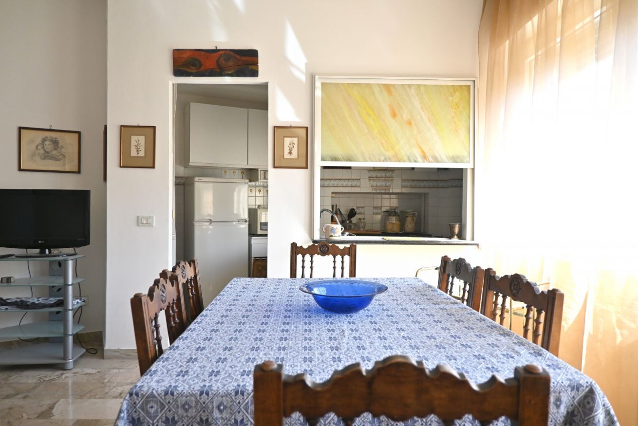 A dining area with a table set for four, a television on a stand, and a kitchen visible through an open door.