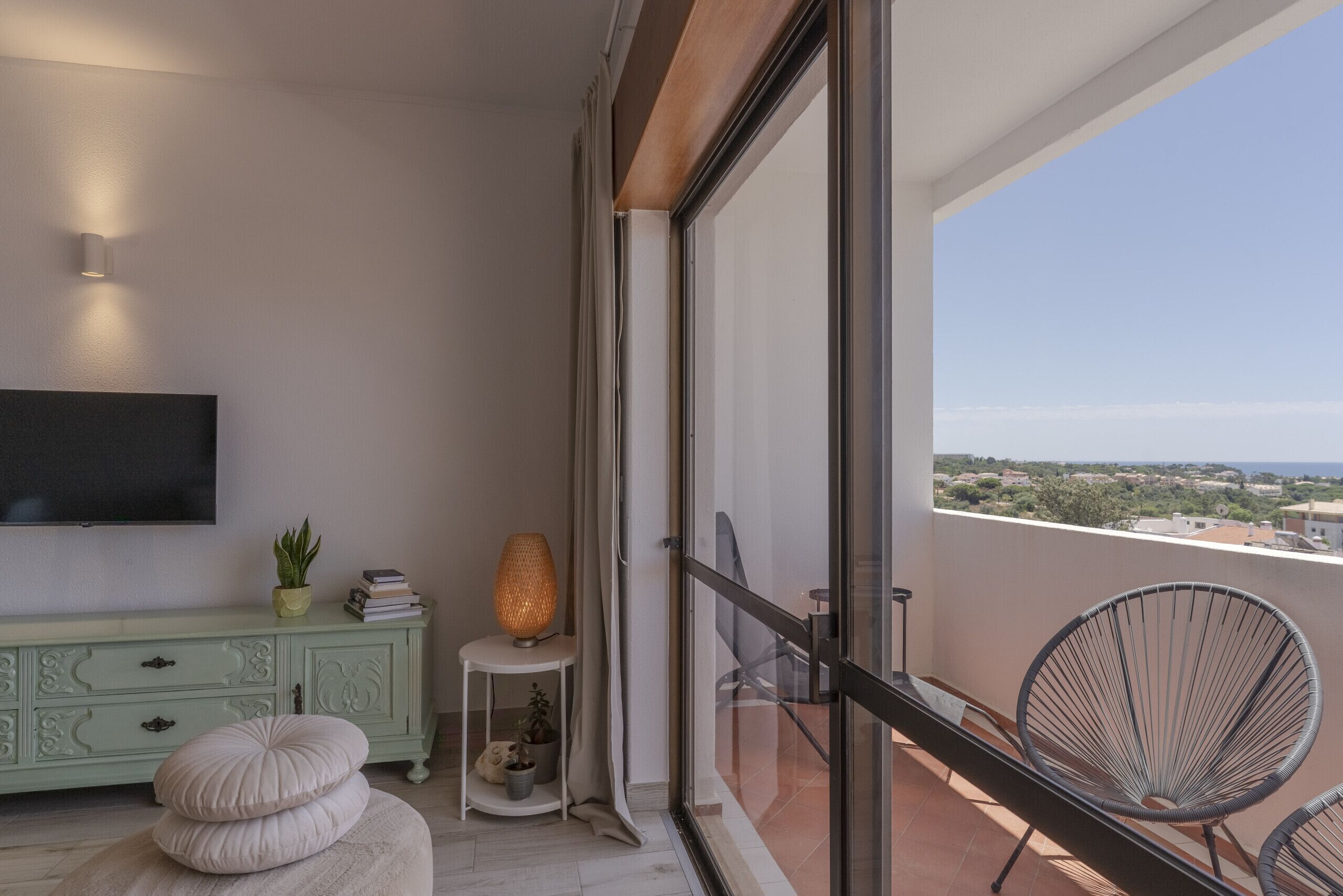 A balcony with a view of the landscape, a television mounted on the wall, a cabinet with decorative items, and a chair.