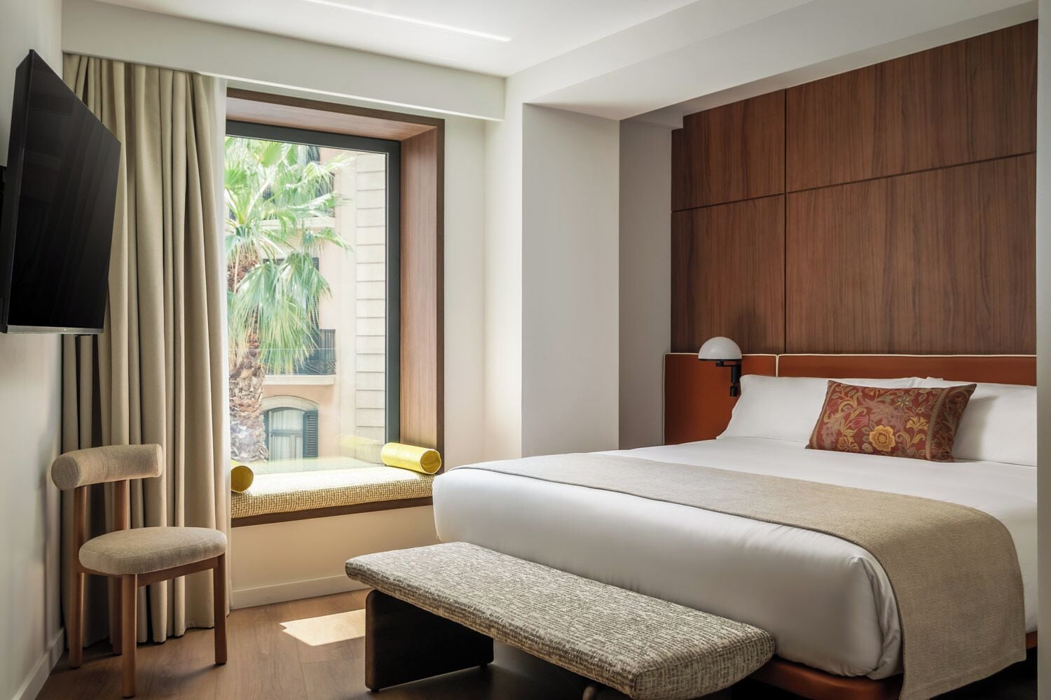 Ein Hotelzimmer mit einem großen Bett, einer Sitzbank, einem Fernseher und einem Fenster mit Vorhängen.