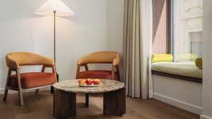 A modern room with two armchairs, a round table with fruit, a floor lamp, and a window with a view of a building. - Casa Aroa Hotel (Barcelona)