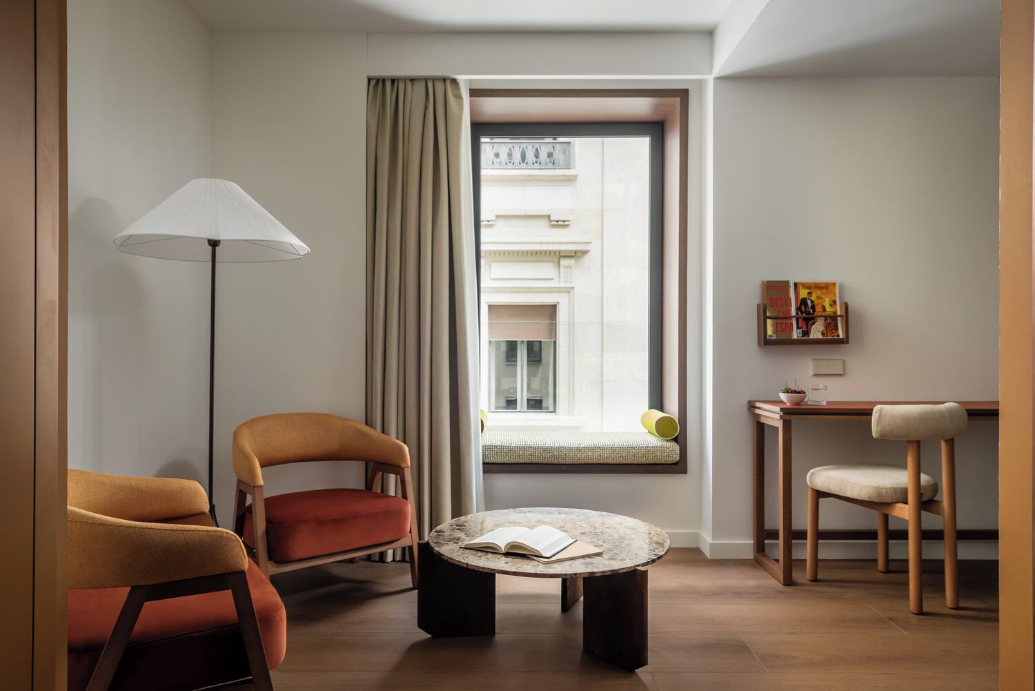 A modern hotel room with a round table, two armchairs, a floor lamp, a window with curtains, and a small desk with a chair.