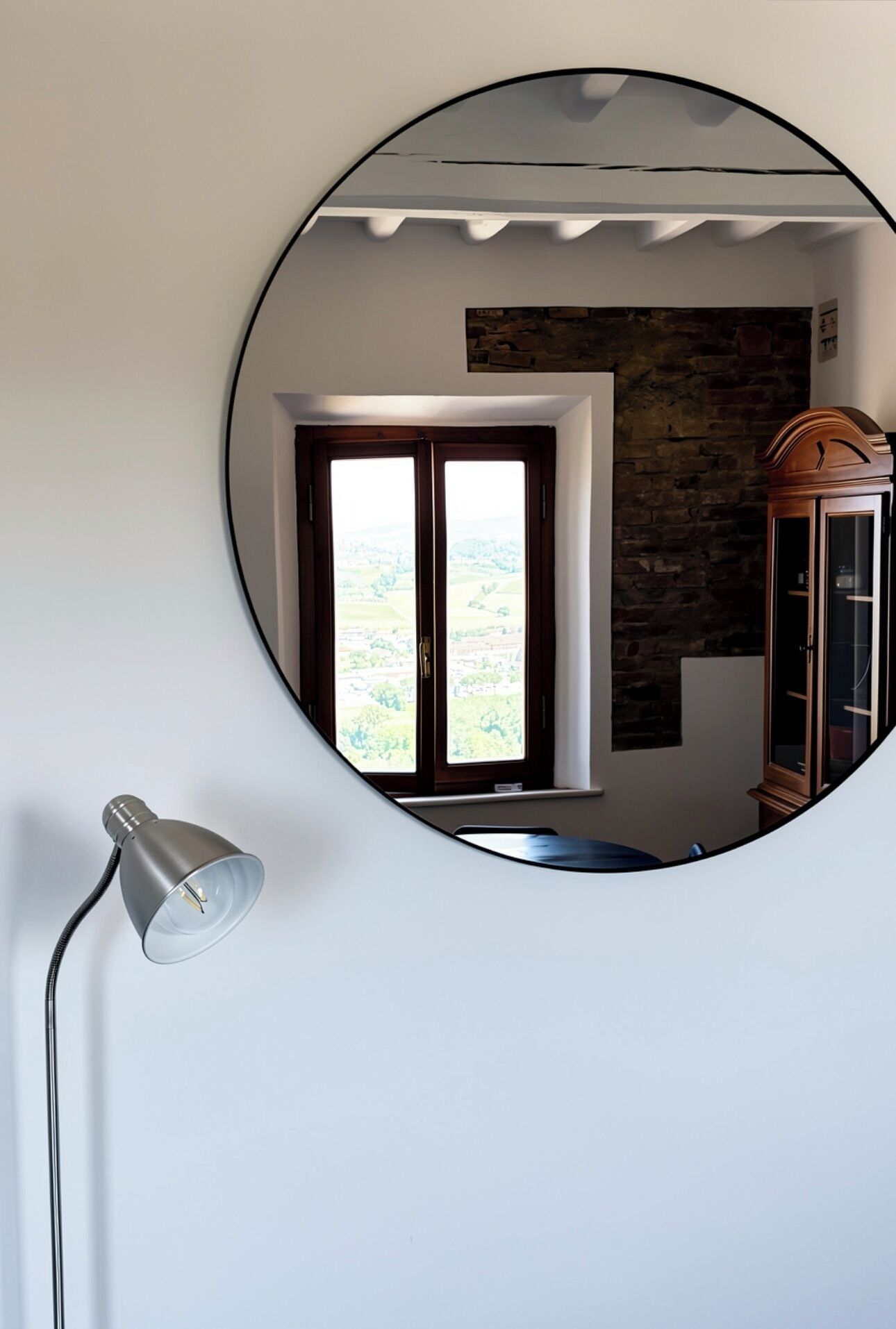 Ein Zimmer mit einem runden Spiegel, der ein Fenster, eine Lampe und einen Holzkasten reflektiert.
