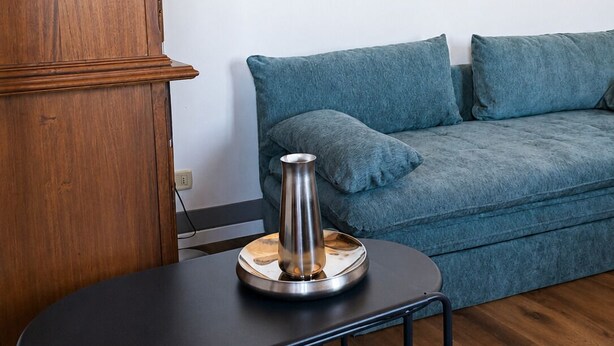 Ein Zimmer mit einem Holzkabinett, einem runden Spiegel an der Wand, einem blauen Sofa und einem kleinen Tisch mit einer Metallvase.