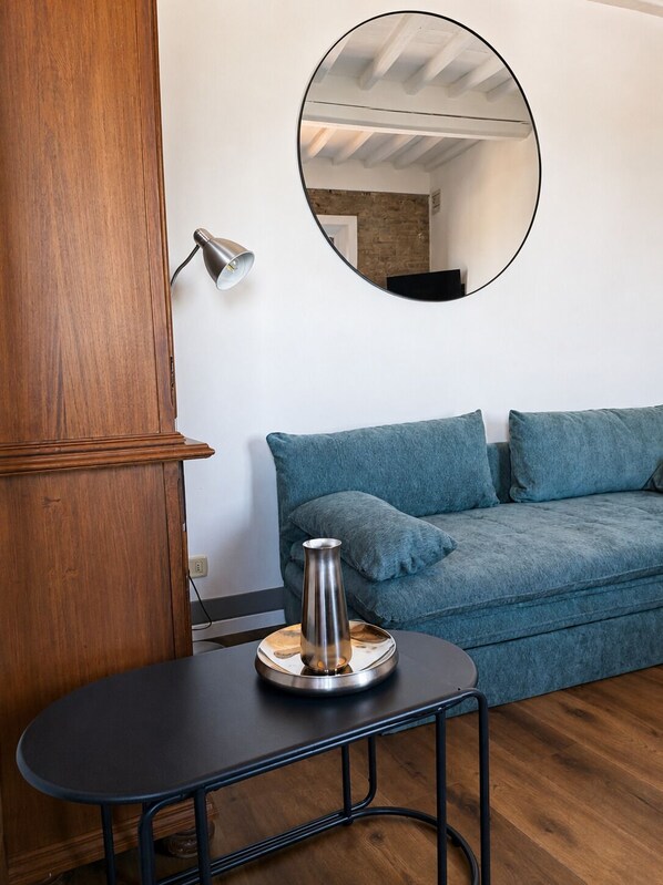 Una habitación con un armario de madera, un espejo redondo en la pared, un sofá azul y una pequeña mesa con un florero metálico.