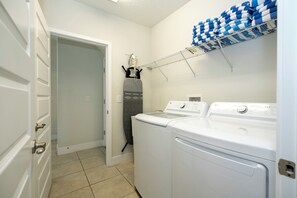 A laundry room with two white front-loading washers and dryers, a drying rack with blue and white towels, and a wall-mounted steam iron. - Luxury Disney Escape - Pool, SPA and Games (Kissimmee)