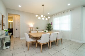 A modern dining room with a wooden table and white chairs, a pendant light, and a view of the outside through a window. - Luxury Disney Escape - Pool, SPA and Games (Kissimmee)