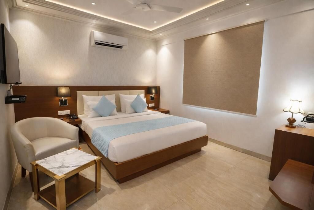 A hotel room with a bed, bedside tables, a chair, a small table, an air conditioning unit, and a television.