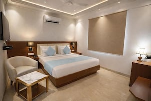 A hotel room with a bed, bedside tables, a chair, a small table, an air conditioning unit, and a television.
