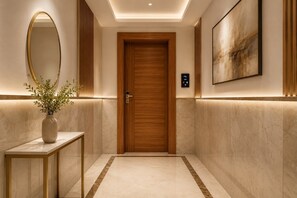 A hallway with a wooden door, a wall-mounted mirror, a vase with plants, and a framed artwork. - NH Skye Suites Hotel T-3 Delhi Airport  (New Delhi)