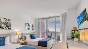 A modern hotel room with a bed, bedside table, lamp, artwork, and a large window with a city view. - Upper Studio Intracoastal & Poolside restaurant (Hallandale Beach)
