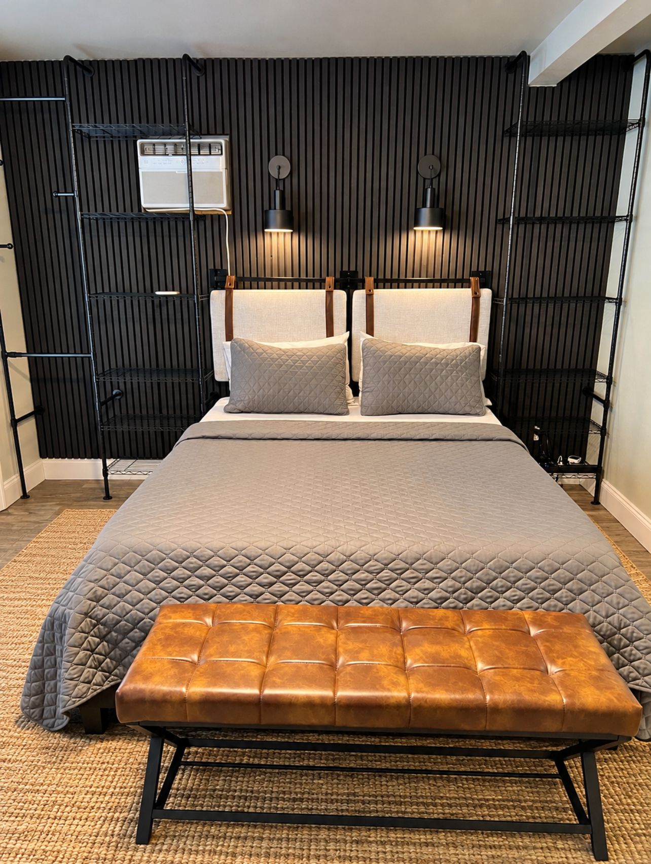 A bed with a quilted cover, two pillows, a bench, and a wall with vertical slats and two wall-mounted lamps.