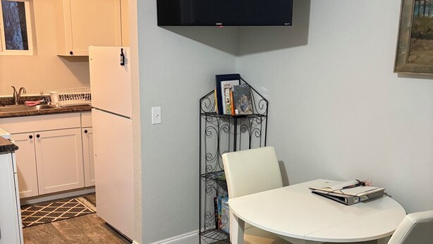 A compact kitchen with white cabinets, a refrigerator, and a dining area with a table and chairs. A flat-screen TV is mounted on the wall above the dining table.