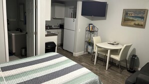 A compact hotel room with a bed, a small kitchenette, a dining area with a table and chairs, and a flat-screen TV mounted on the wall.