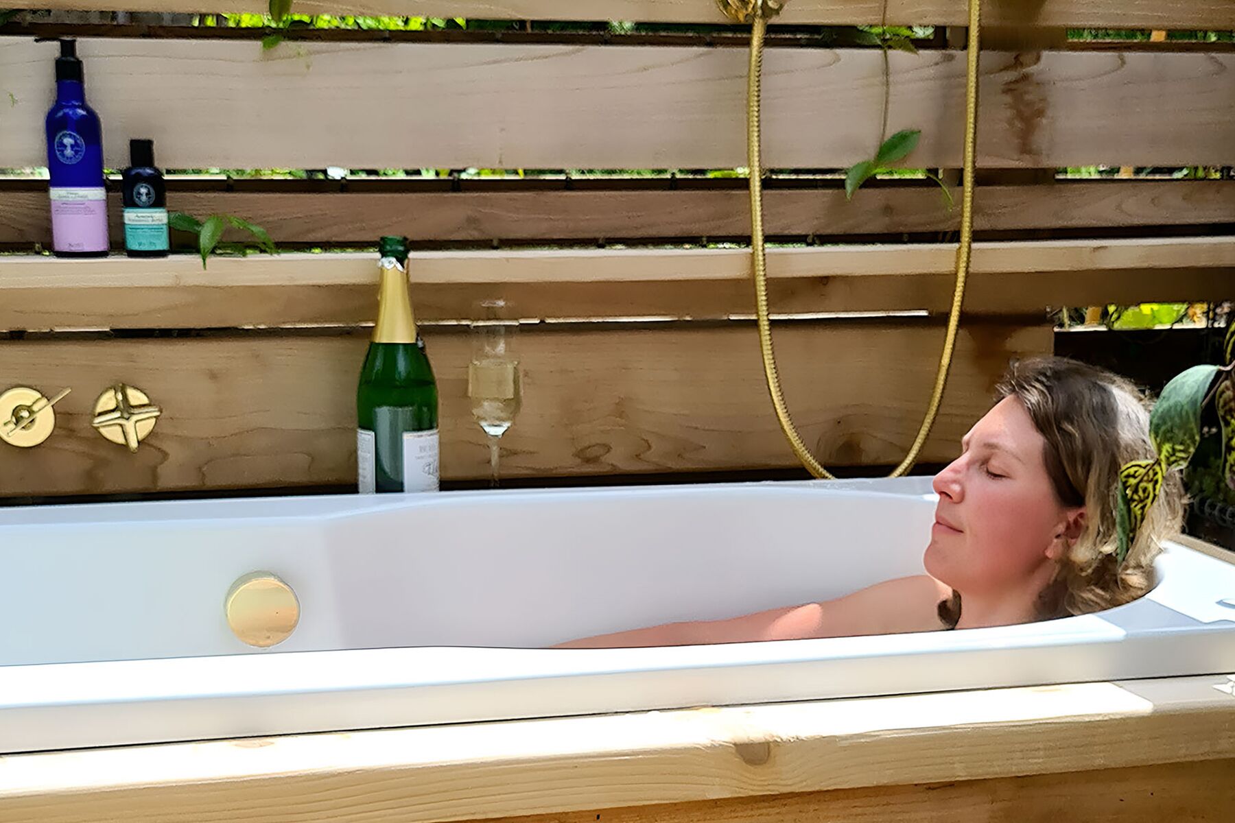 A person relaxing in a bathtub with a bottle of champagne and a glass of wine on a wooden shelf.