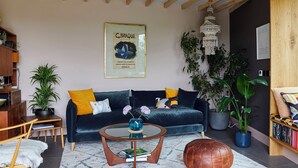 A living room with a dark blue sofa, a coffee table, a leather pouf, and a hanging light fixture. - Award-winning Nuthatch Retreat | Cinema | Landscaped gardens | Bathe under stars (Blakeney)