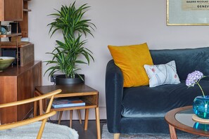 Ein Wohnzimmer mit einem dunklen Sofa, einem gelben Kissen, einer blauen Vase mit Blumen, einem Holztischchen, einer Topfpflanze und einem Bücherregal.