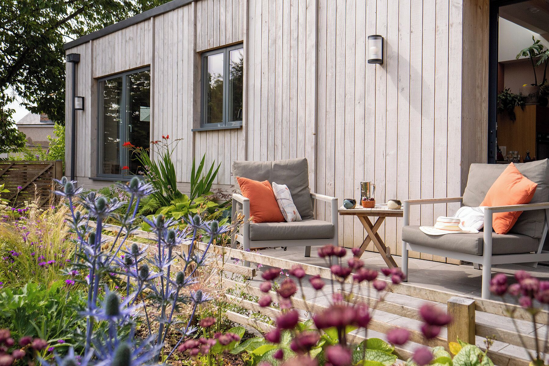 Une maison moderne en bois, avec deux canapés gris et une petite table, entourée d’un jardin où poussent des fleurs violettes.