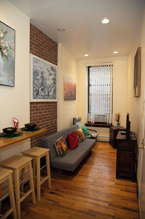 A living room with a sofa, a dining table with stools, a brick wall, and artwork on the walls.
