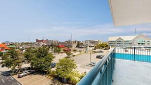 A view from a balcony overlooking a parking lot with several cars, a swimming pool, and buildings in the background. - Sunset Pointe 302 - Pool, Prime Location (Ocean City)