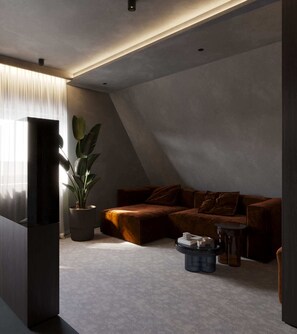 Ein modernes Wohnzimmer mit schrägverdeckter Decke, einem dunkelgrauen Sofa, einer Topfpflanze und einem kleinen runden Tisch.