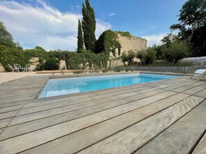 Una piscina rectangular amb un tram de fusta al voltant, una paret de pedra i un arbre alt al fons.