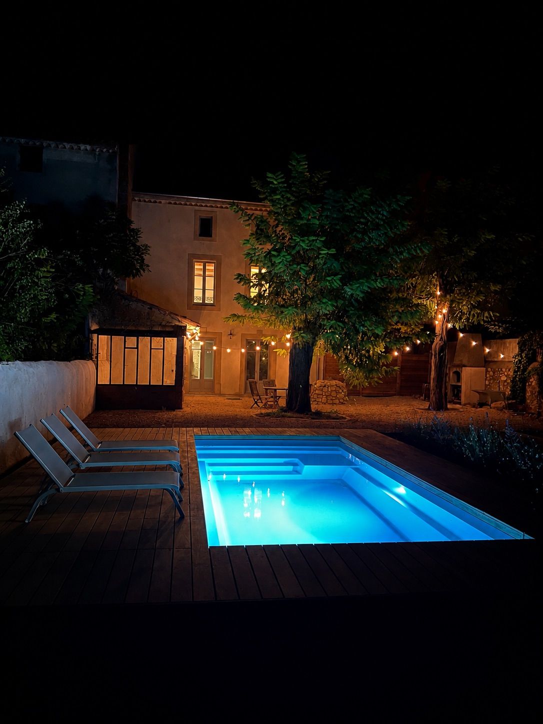 Une piscine avec une terrasse en bois et des chaises longues, entourée d’une maison décorée de guirlandes lumineuses et bord ée d’arbres.