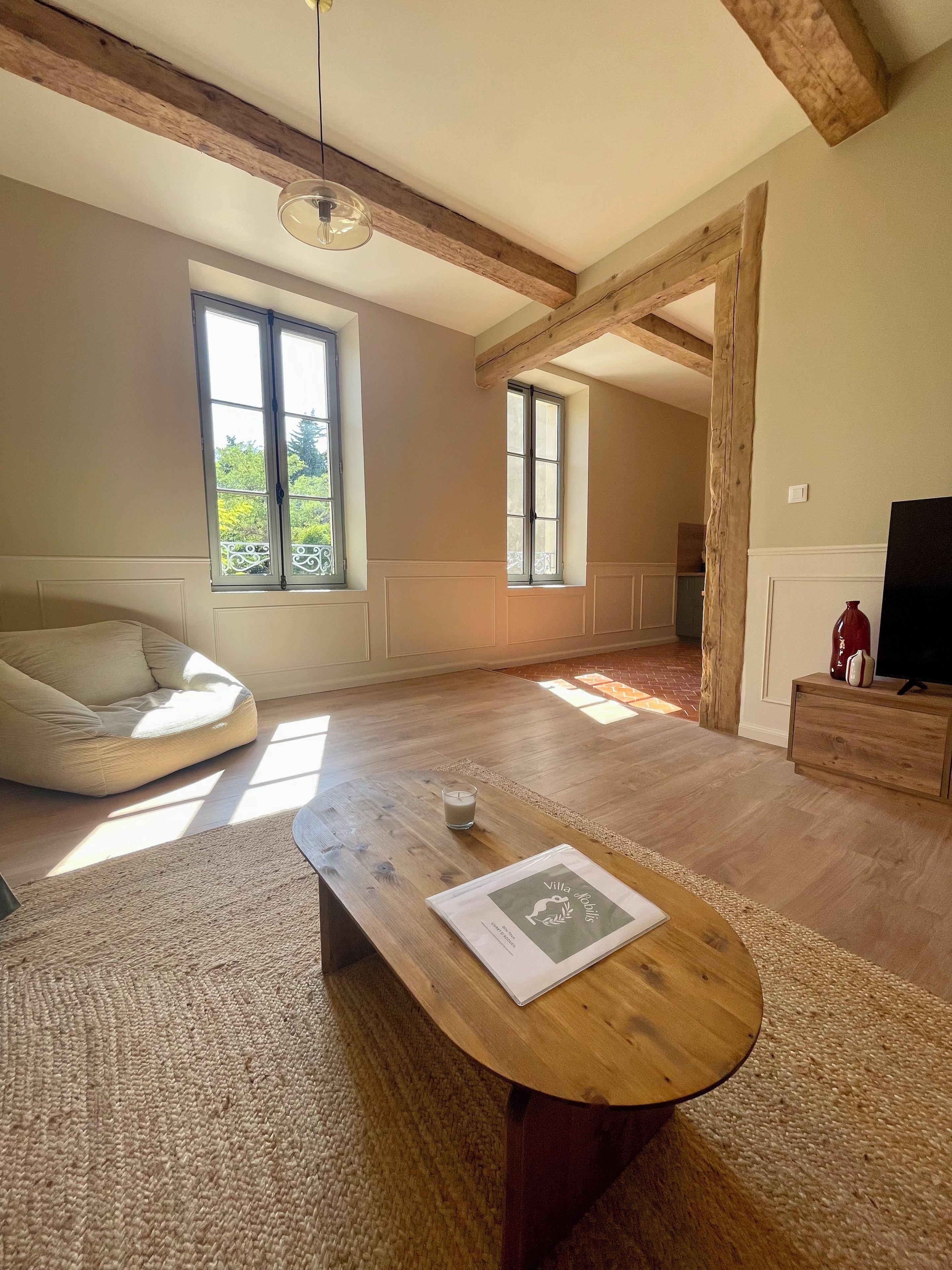 A spacious living room with a wooden floor, a round coffee table, a bean bag chair, and large windows allowing natural light.